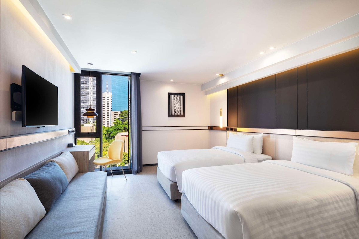 A modern hotel room with two beds, a sofa, a flat-screen TV, and a large window offering a city view.