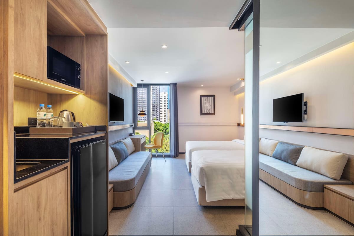 A modern hotel room with a kitchenette, a sofa bed, a TV, and a large window offering a city view.