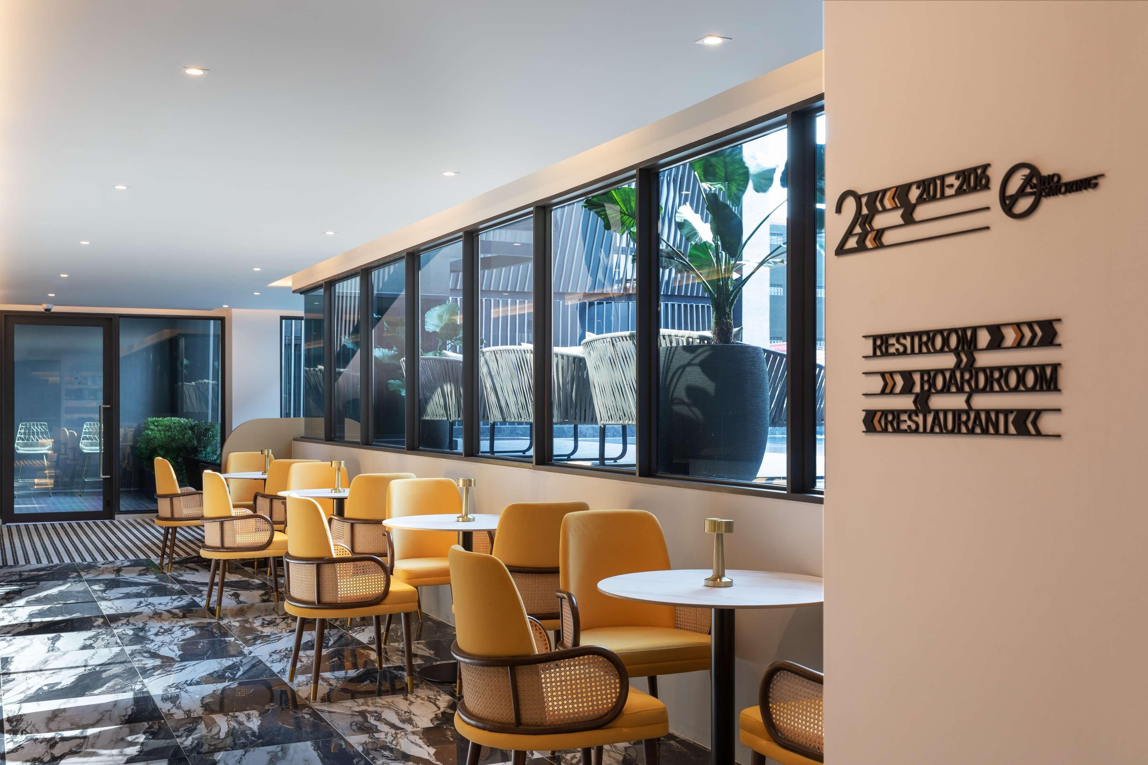 Un interior moderno con sillas y mesas amarillas, ventanas amplias y un cartel que indica servicios como un baño, sala de reuniones y restaurante.