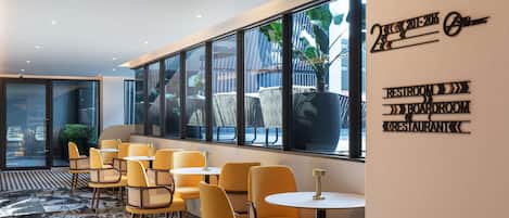 Un interior moderno con sillas y mesas amarillas, ventanas amplias y un cartel que indica servicios como un baño, sala de reuniones y restaurante.