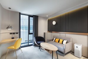 Et moderne hotelværelse med sofa, stol, lille bord, vægur og et vindue med gardiner.
