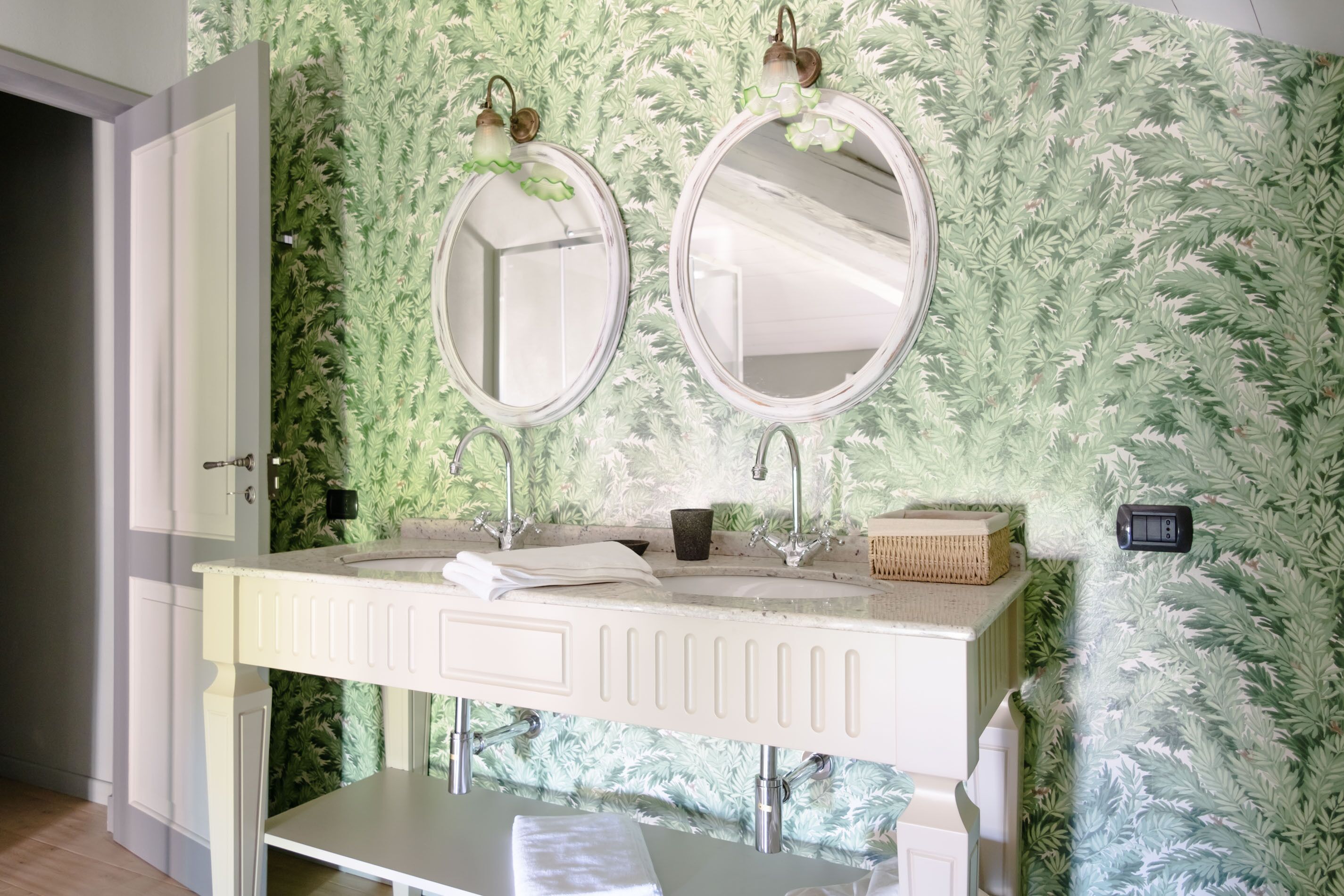 A bathroom with a white vanity, two round mirrors, a sink, and a green leaf-patterned wall.