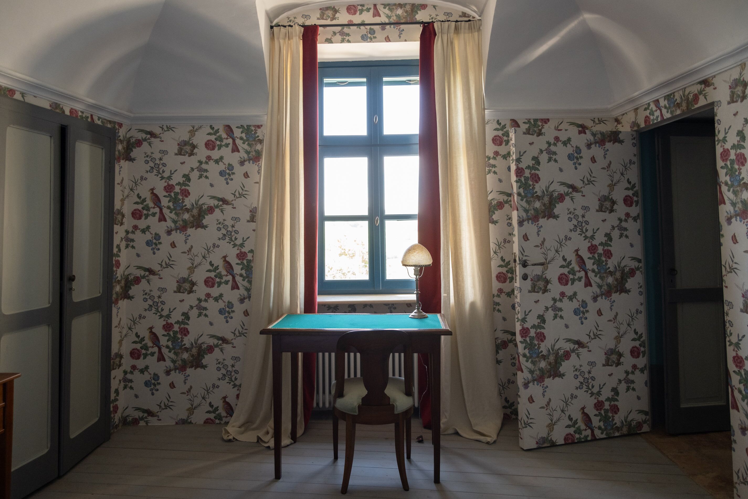 Una habitación con papel tapiz floral, una ventana con cortinas rojas, una mesa con lámpara y una silla.