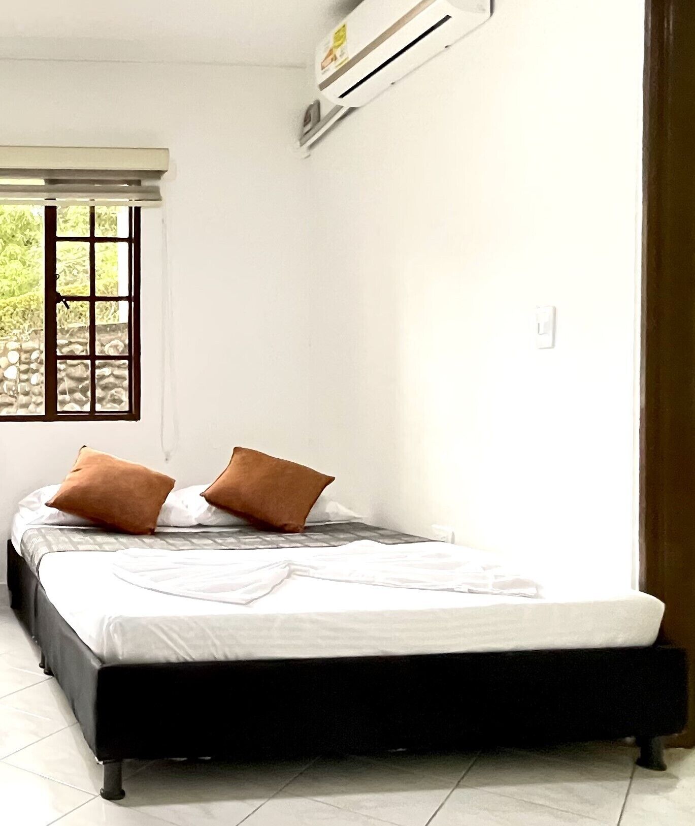 A single bed with a black frame, white bedding, and two brown pillows. There is a window with a view of greenery outside.