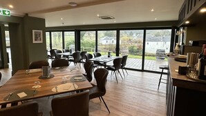 Un vaste espace dédié à la salle à manger, avec une longue table en bois et de nombreuses chaises, de grandes fenêtres offrant une vue sur l’extérieur, et un bar avec divers articles exposés sur le comptoir.