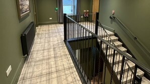Un couloir avec un tapis à carreaux, un radiateur, un tableau encadré accroché au mur et un escalier doté d’une rampe en métal.