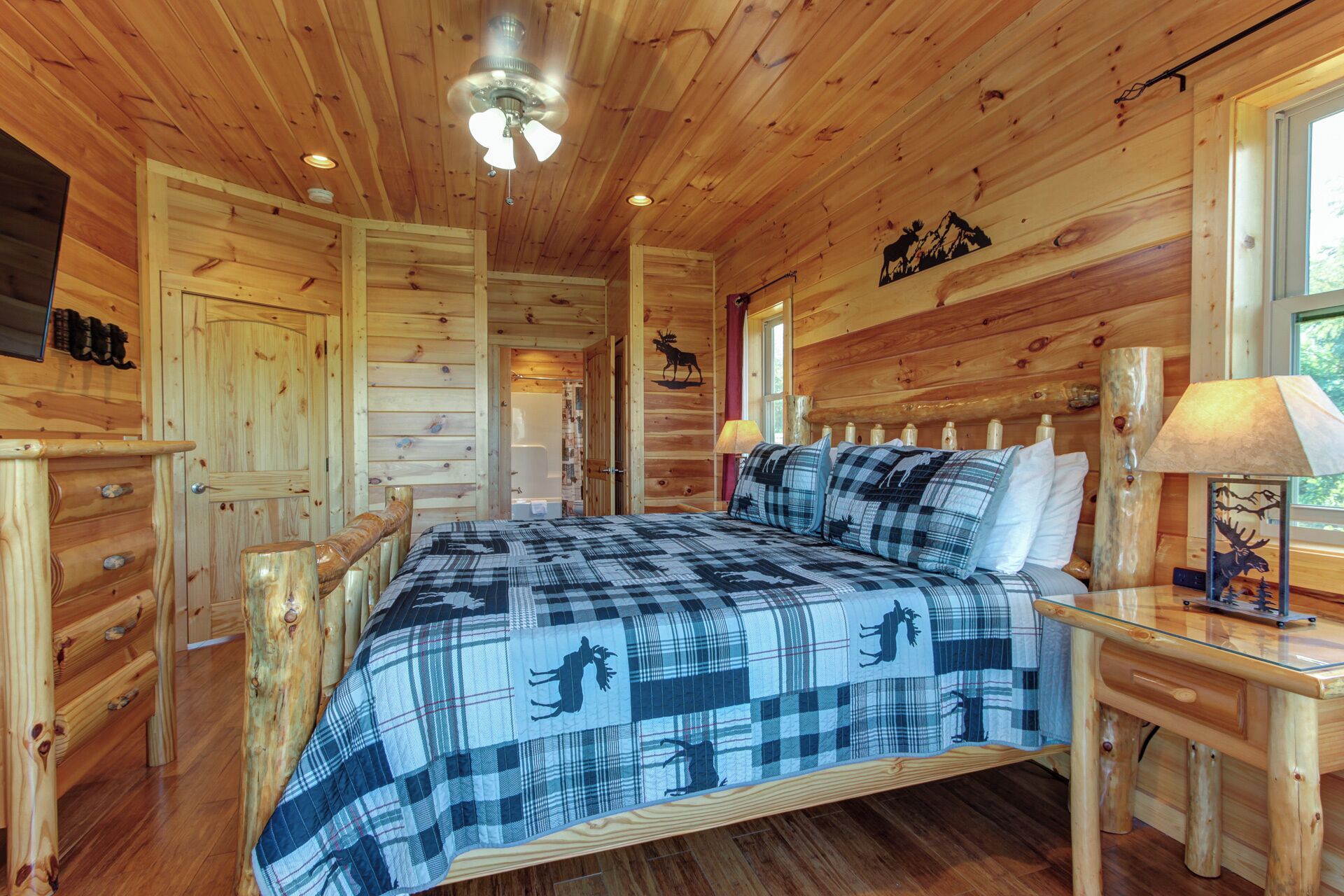 A wooden cabin room with a bed, a wooden dresser, a nightstand, a lamp, and a ceiling fan.