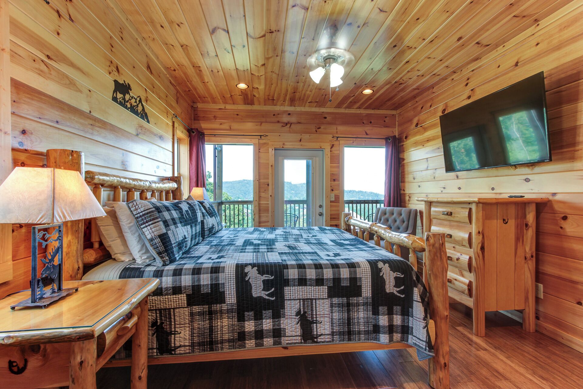 A wooden cabin bedroom with a bed, a TV, a desk, and a balcony with a view of the mountains.