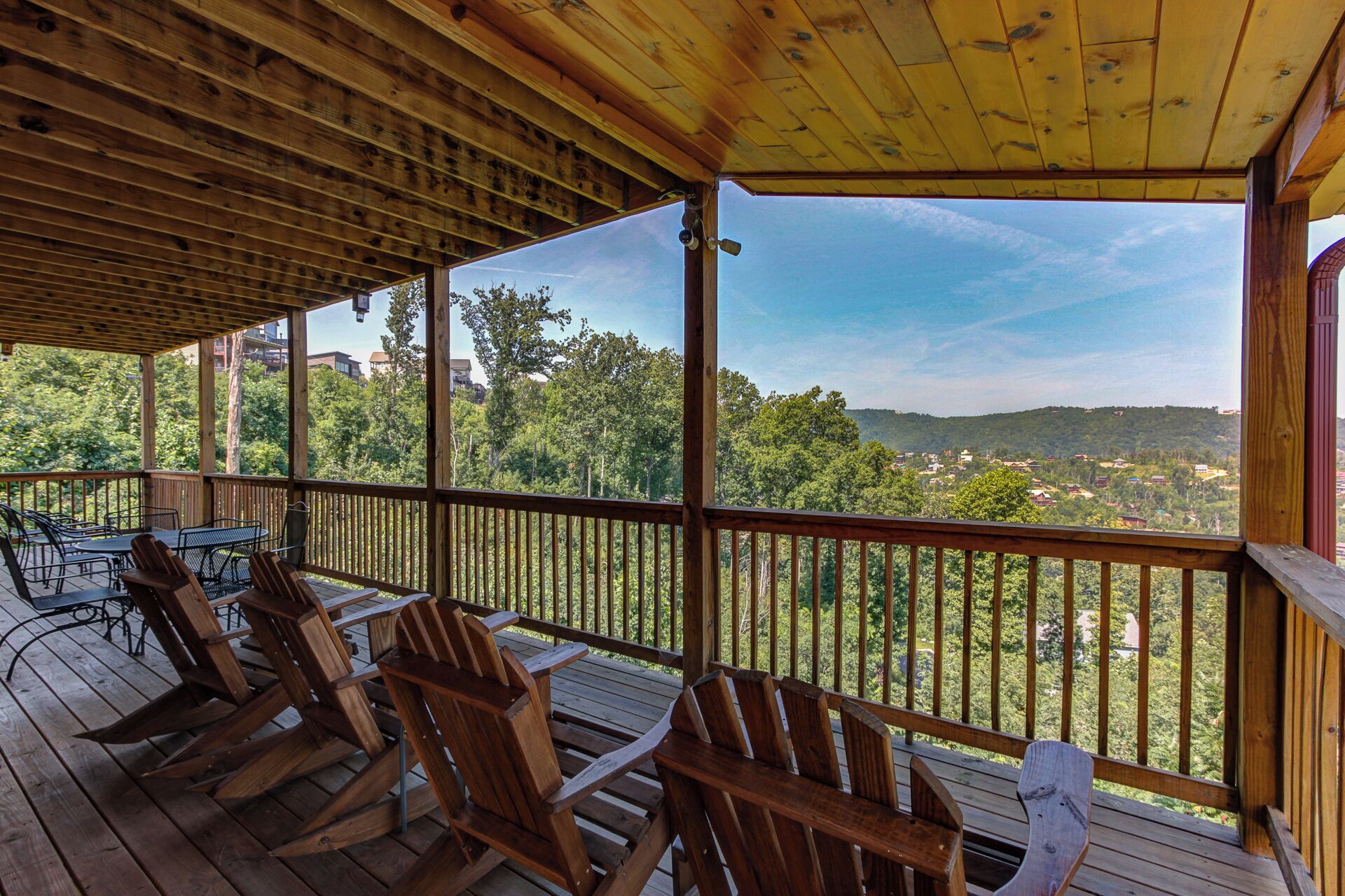 Une terrasse en bois avec des fauteuils Adirondack, une table et une vue sur une zone boisée, avec des bâtiments visibles au loin.