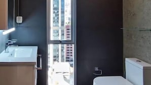 Une salle de bain moderne équipée de toilettes, d’un lavabo et d’un miroir. Une fenêtre offre une vue sur les bâtiments environnants.