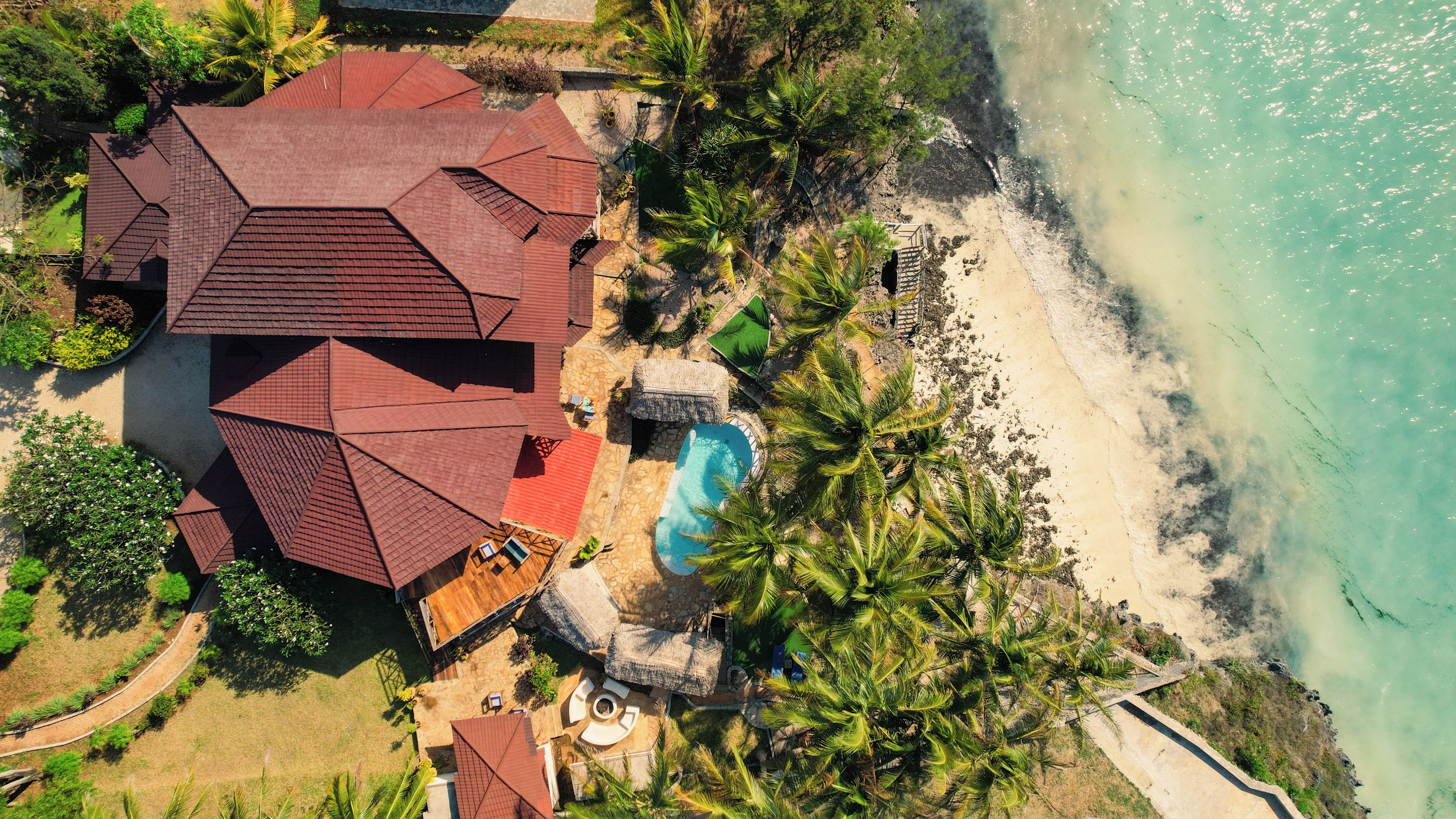 An aerial view of a coastal property with a swimming pool, a thatched-roof structure, and a beach.