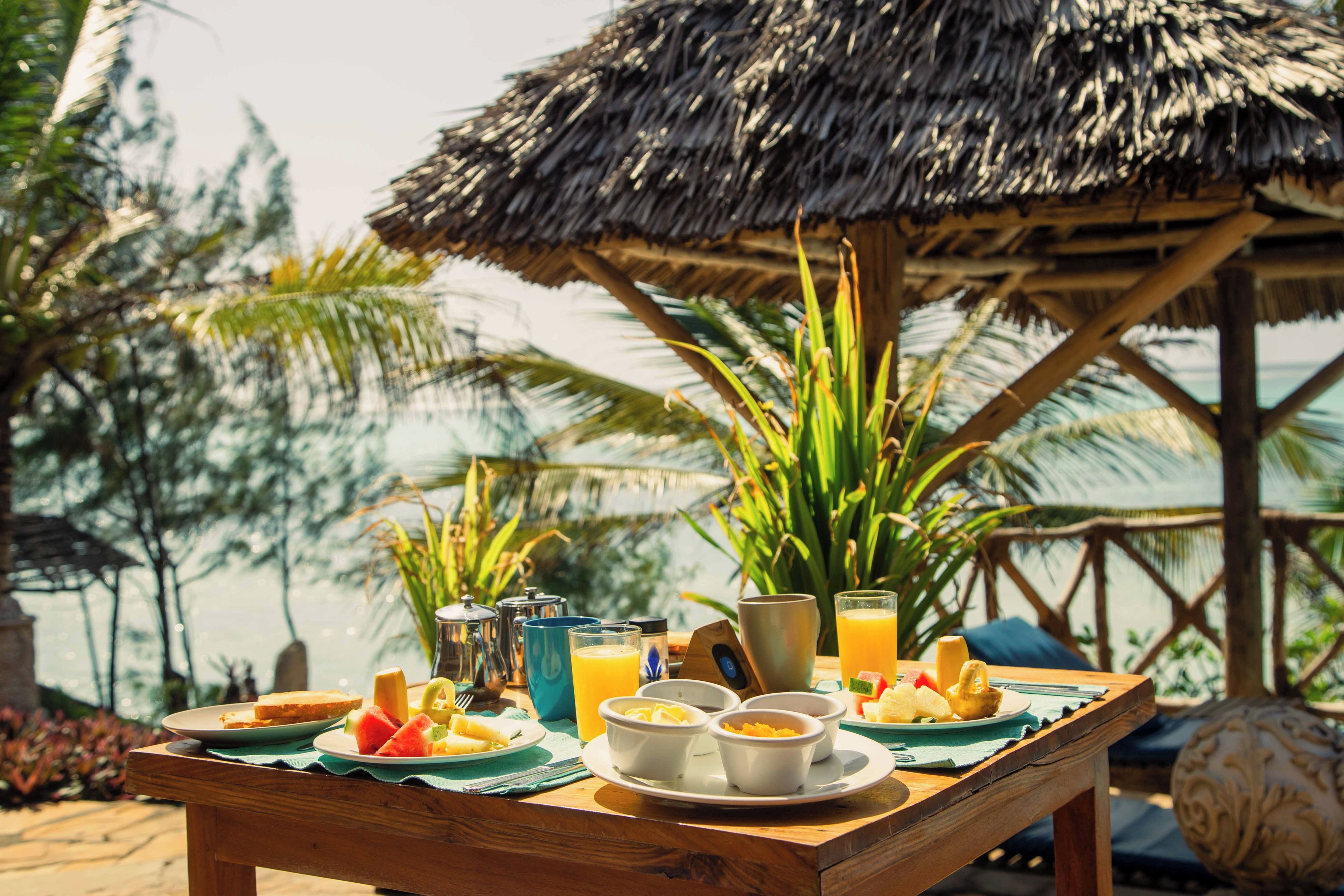 Una mesa de madera con alimentos para el desayuno, como tostadas, fruta y jugo de naranja, ubicada al aire libre con un techo de paja y palmeras al fondo.