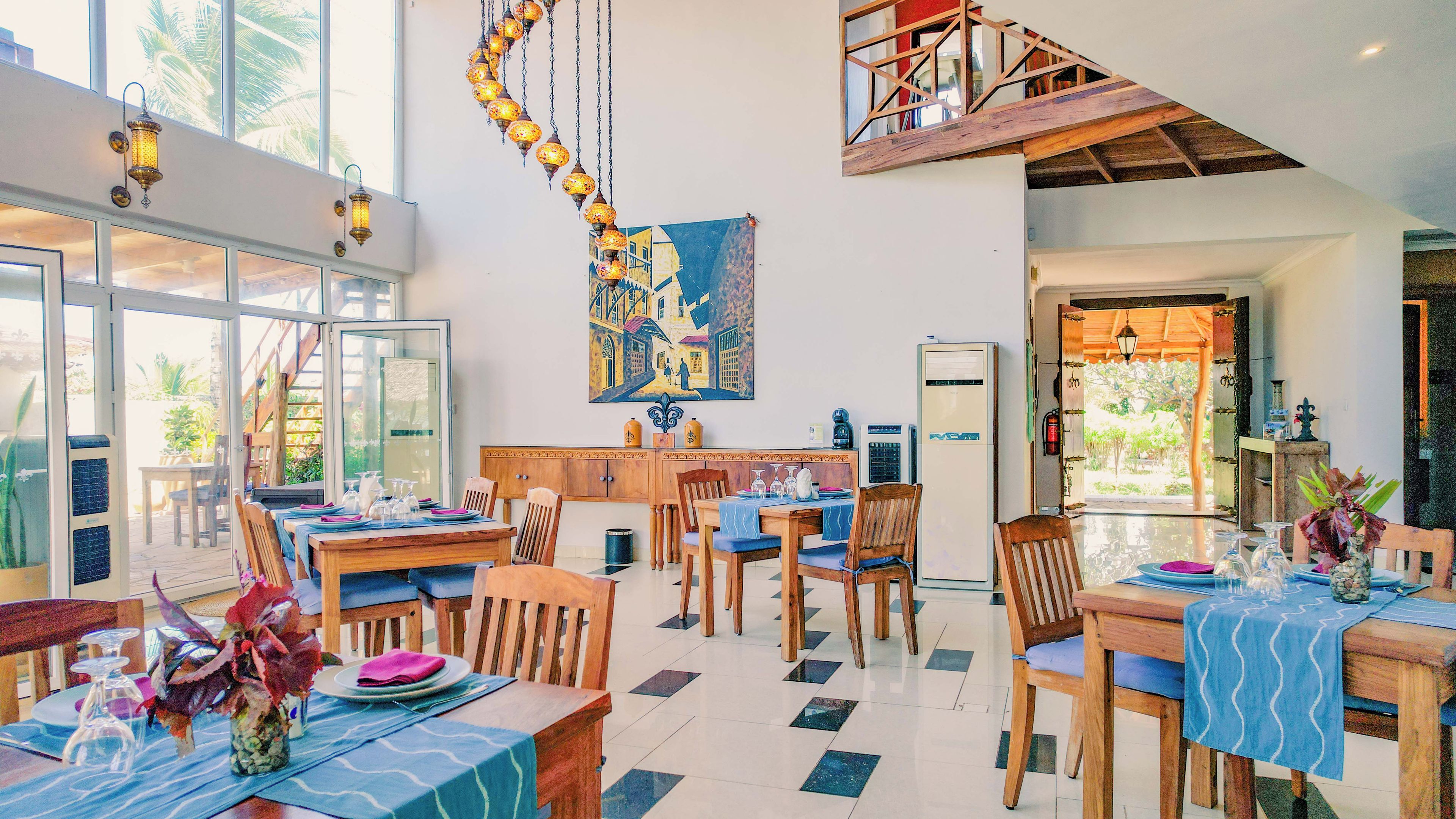 A dining area with multiple tables and chairs, a kitchen area with a refrigerator, and a staircase leading to an upper level.