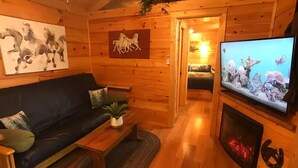 L’intérieur d’un chalet accueillant, avec un plancher en bois, un canapé noir agrémenté de coussins, une table basse en bois, une cheminée surmontée d’un téléviseur et un tableau représentant des chevaux accroché au mur.
