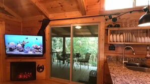 L’intérieur d’un chalet en bois comprend une télévision, une cheminée, des portes-fenêtres coulissantes donnant sur une terrasse et un coin cuisine.