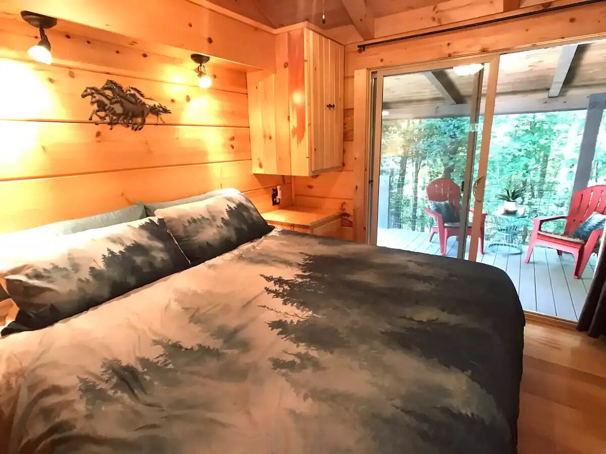 A wooden cabin bedroom with a bed, a nightstand, and a sliding glass door leading to an outdoor deck with chairs.