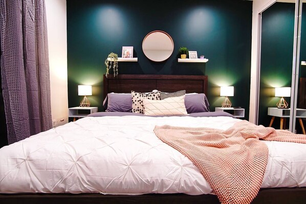 A bedroom with a large bed, dark wooden headboard, two bedside tables with lamps, a round mirror, and a green accent wall. - Beach Breeze Bungalow King (Lawndale)