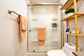 A modern bathroom with a glass-enclosed shower, a toilet, and a shelf with toiletries. - Beach Breeze Bungalow King (Lawndale)