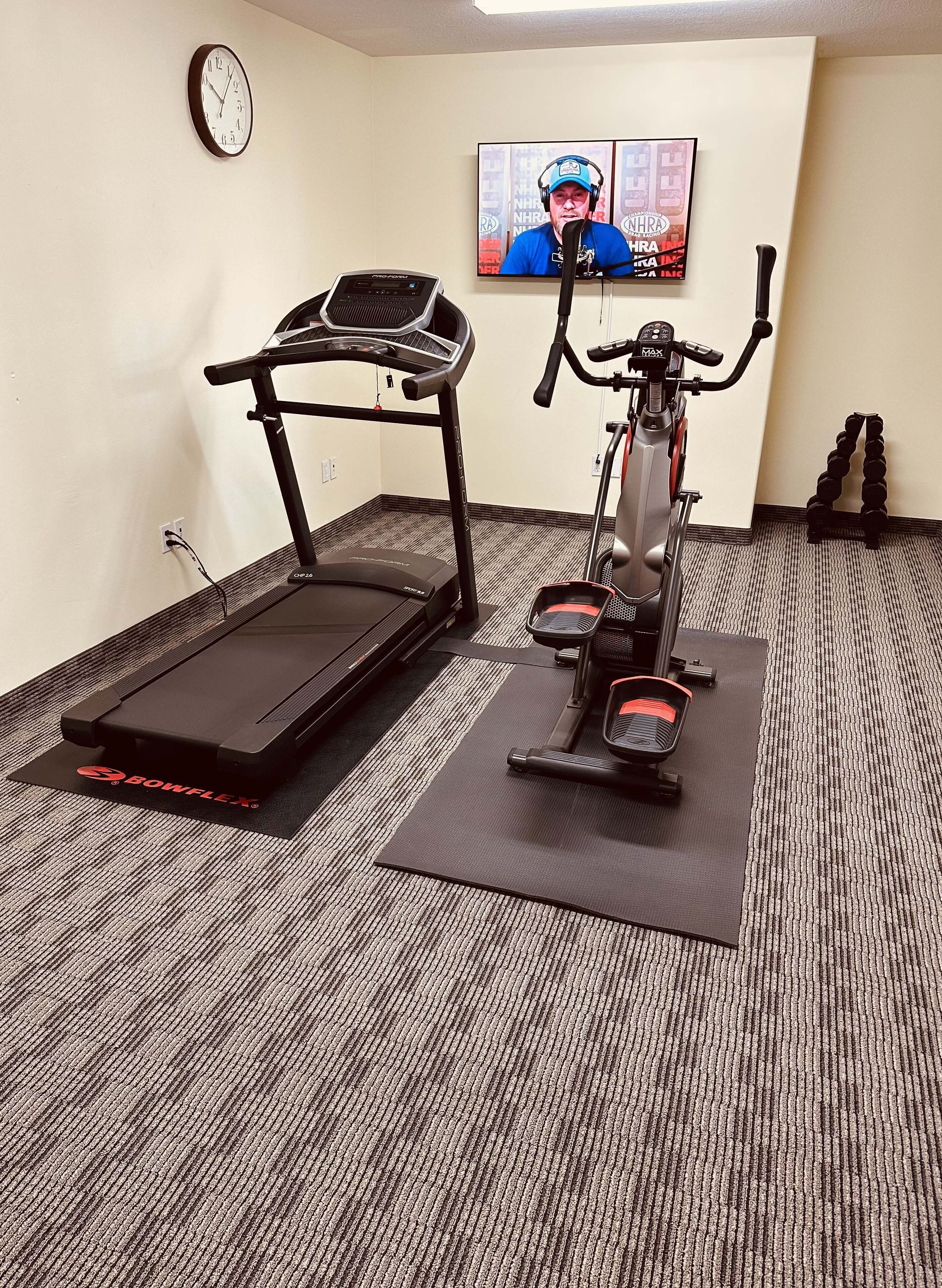 A home gym with a treadmill and an elliptical machine, a clock on the wall, and a TV displaying a sports event.