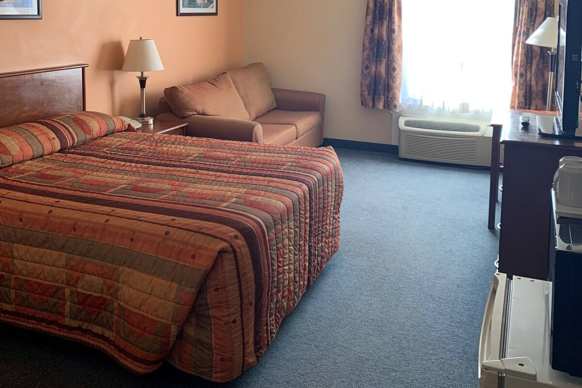 Ein Hotelzimmer mit Bett, Schreibtisch, Stuhl, Lampe, einem Bild an der Wand und einem Vorhangfenster.