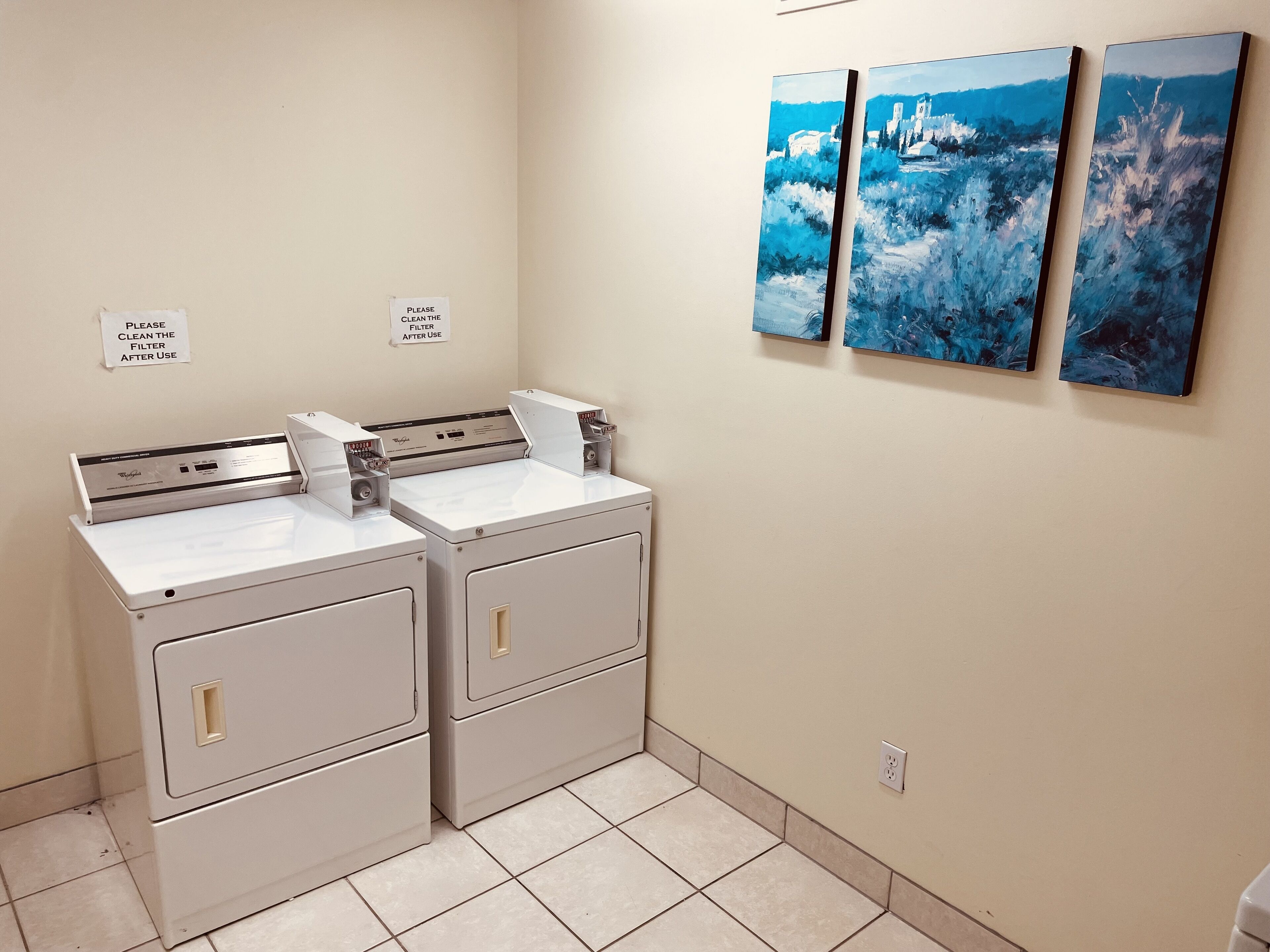 Une buanderie équipée de deux machines à laver et de deux sèche-linge à chargement frontal, d’un panneau fixé au mur indiquant qu’il faut nettoyer le filtre après chaque utilisation, et de trois tableaux abstraits accrochés au mur.