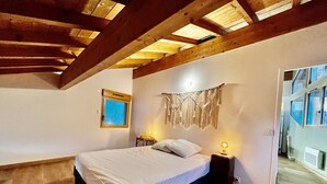 Una habitació amb sostre de fusta, un llit amb roba de llit blanca, una taula de nit amb una làmpada i una finestra amb vista.