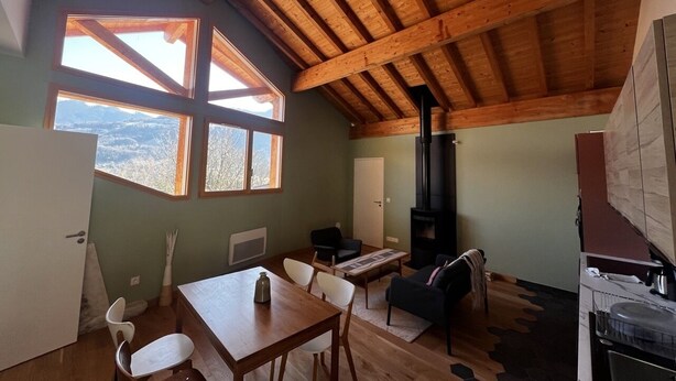 Una habitació acollidora amb sostre de fusta, una llar de foc, una taula de menjador amb caires i grans finestres que donen a l'exterior.