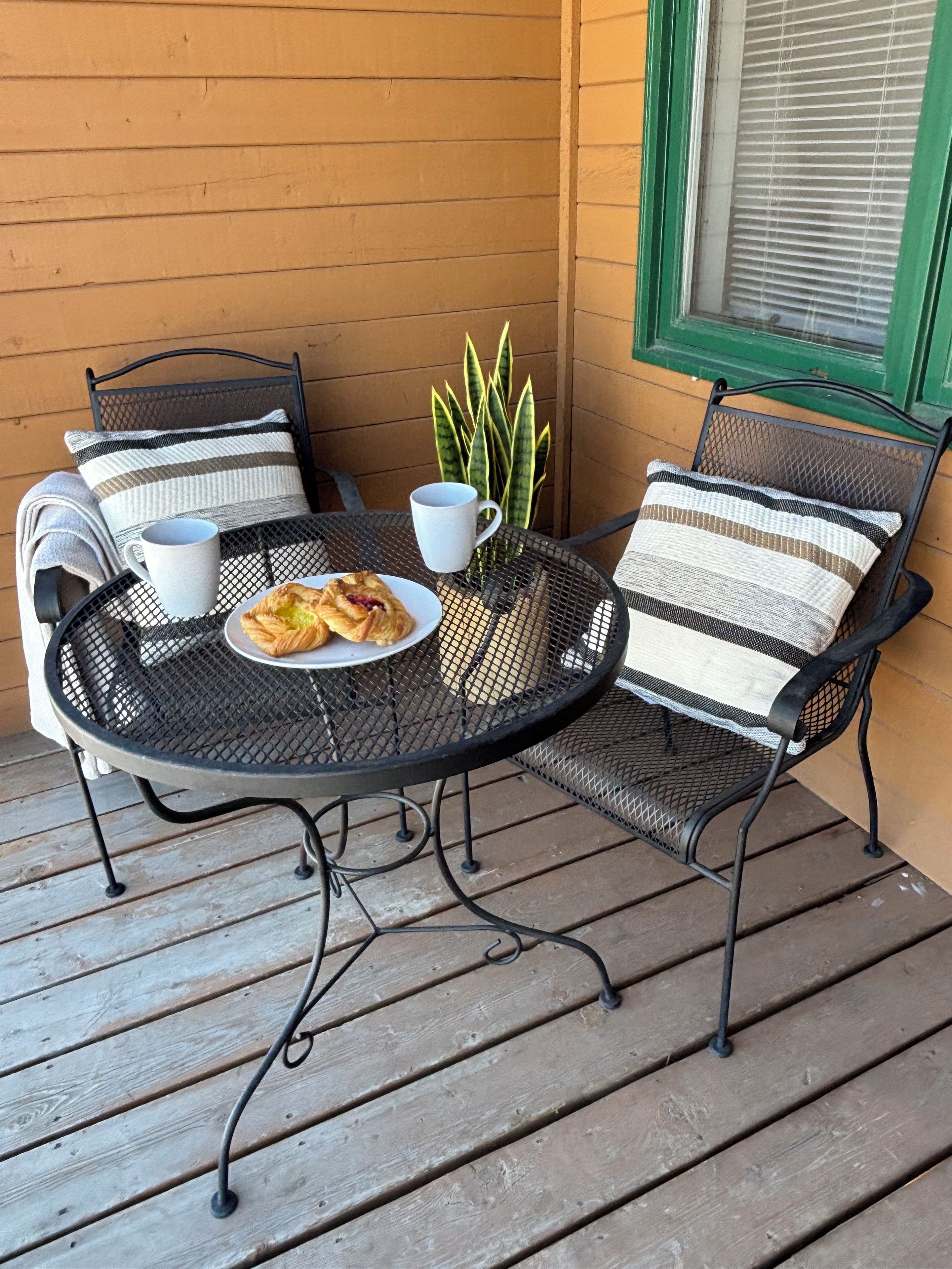 A round metal table with two chairs, a plate of food, and two cups on a wooden deck.