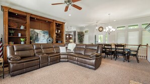 A spacious living room with a large leather sectional sofa, wooden built-in shelving, a dining area with a wooden table and chairs, and a ceiling fan. - Overlooking 9th Green at Osage National Golf Resort (Lake Ozark)