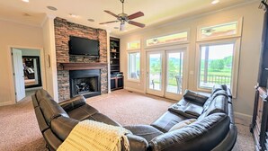 A living room with a stone fireplace, a flat-screen TV, a ceiling fan, and large windows offering a view of the outdoors. - Overlooking 9th Green at Osage National Golf Resort (Lake Ozark)
