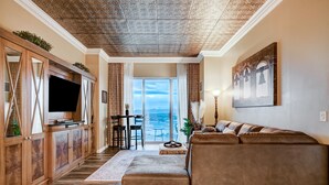 A living room with a large flat-screen TV, wooden cabinets, a brown leather sofa, and a view of the ocean through a glass door.