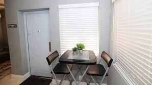 Un pequeño comedor con una mesa y cuatro sillas, una puerta blanca y un ventanal con persianas.