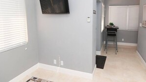 Una habitación con paredes grises, un televisor montado, una mesita con una silla y una ventana con persianas.