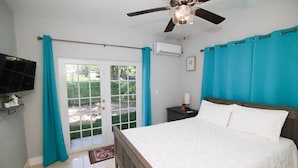 Un dormitorio con cama, ventilador de techo, televisor montado en la pared y puerta que da a un espacio exterior.