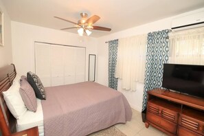 A bedroom with a bed, a ceiling fan, a TV, a wardrobe, and a window with patterned curtains.