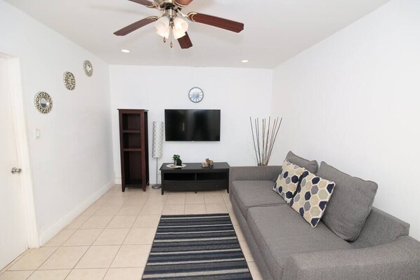 A living room with a grey sofa, a flat-screen TV, a ceiling fan, and a tiled floor.
