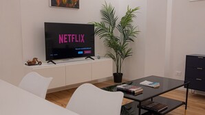 Un salón moderno con un televisor de pantalla plana mostrando 'NETFLIX', una planta en maceta, una mesa de centro de vidrio con libros y un sofá blanco.