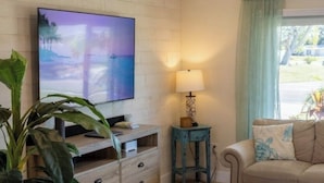 A living room with a flat-screen TV, wooden furniture, a lamp, a sofa, and a window with curtains. - The Sand Castle - Backyard Oasis Mins from Siesta! (Sarasota)