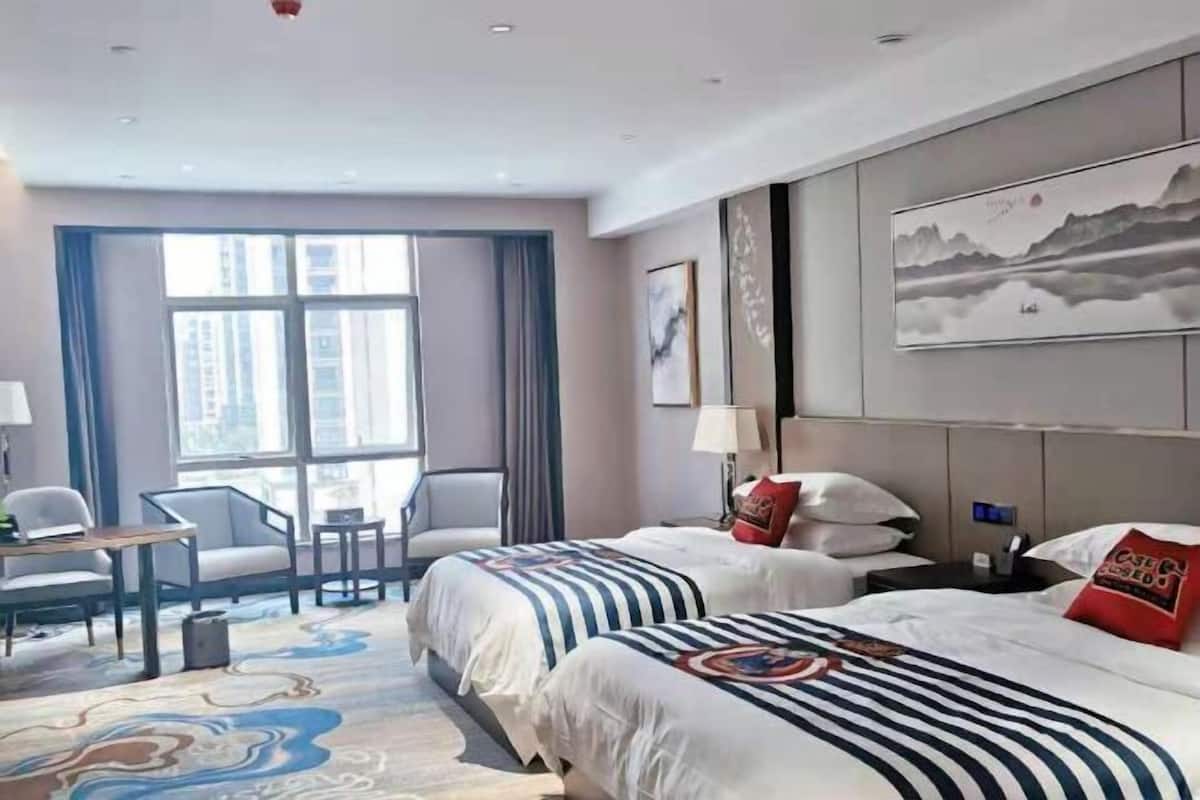 Une chambre d’hôtel avec deux lits, un bureau, des chaises, une grande fenêtre et des œuvres d’art accrochées aux murs.