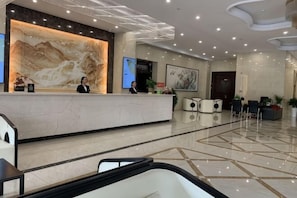 Uma área de recepção de hotel com um balcão de mármore, dois recepcionistas, uma grande obra de arte emoldurada e um chão de azulejos limpo.