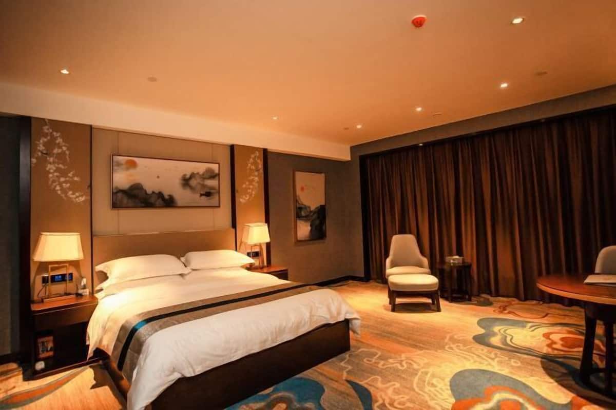 Une chambre d’hôtel avec un grand lit, deux tables de chevet avec des lampes, une chaise, une petite table et une grande fenêtre avec des rideaux.