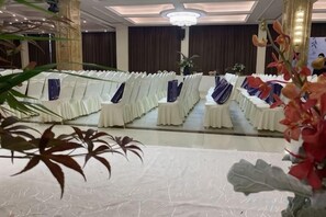 Une salle de banquet avec des rangées de tables et de chaises blanches, un lustre et un grand miroir qui reflète la pièce.