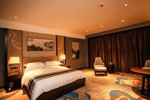Ein Hotelzimmer mit einem großen Bett, zwei Nachttischen, einem Sessel, einem kleinen Tisch und einem großen Fenster mit Vorhängen.