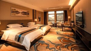 Une chambre d’hôtel avec deux lits, un bureau, des chaises, une grande fenêtre et des œuvres d’art accrochées aux murs.