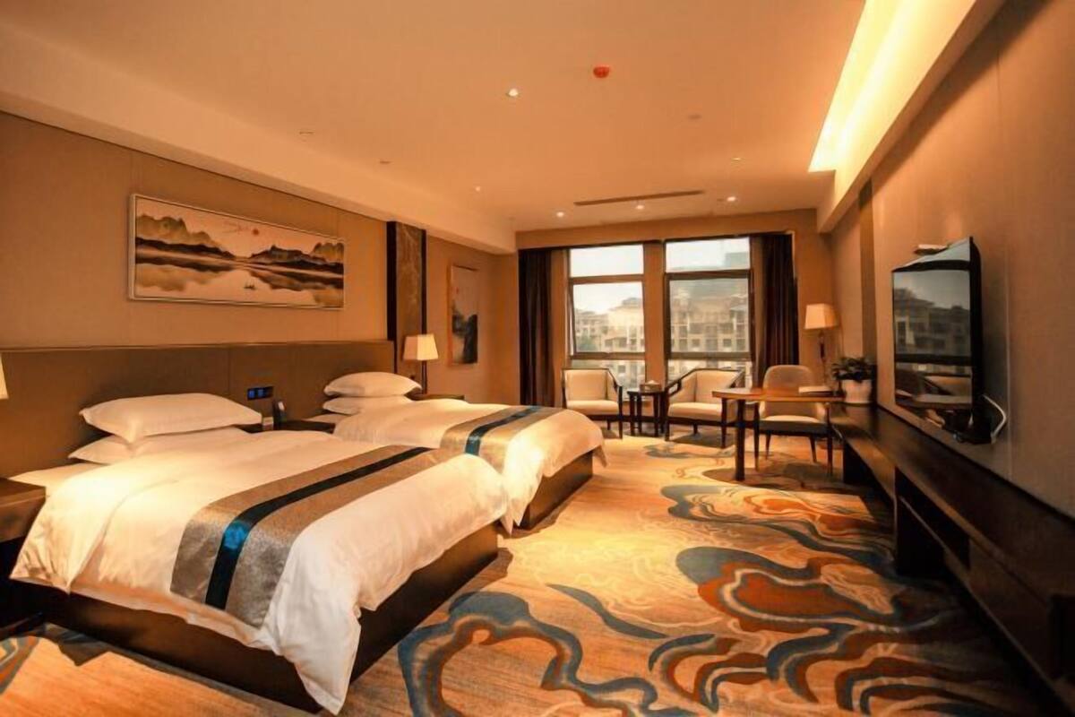 Une chambre d’hôtel avec deux lits, un bureau, des chaises, une grande fenêtre et des œuvres d’art accrochées aux murs.