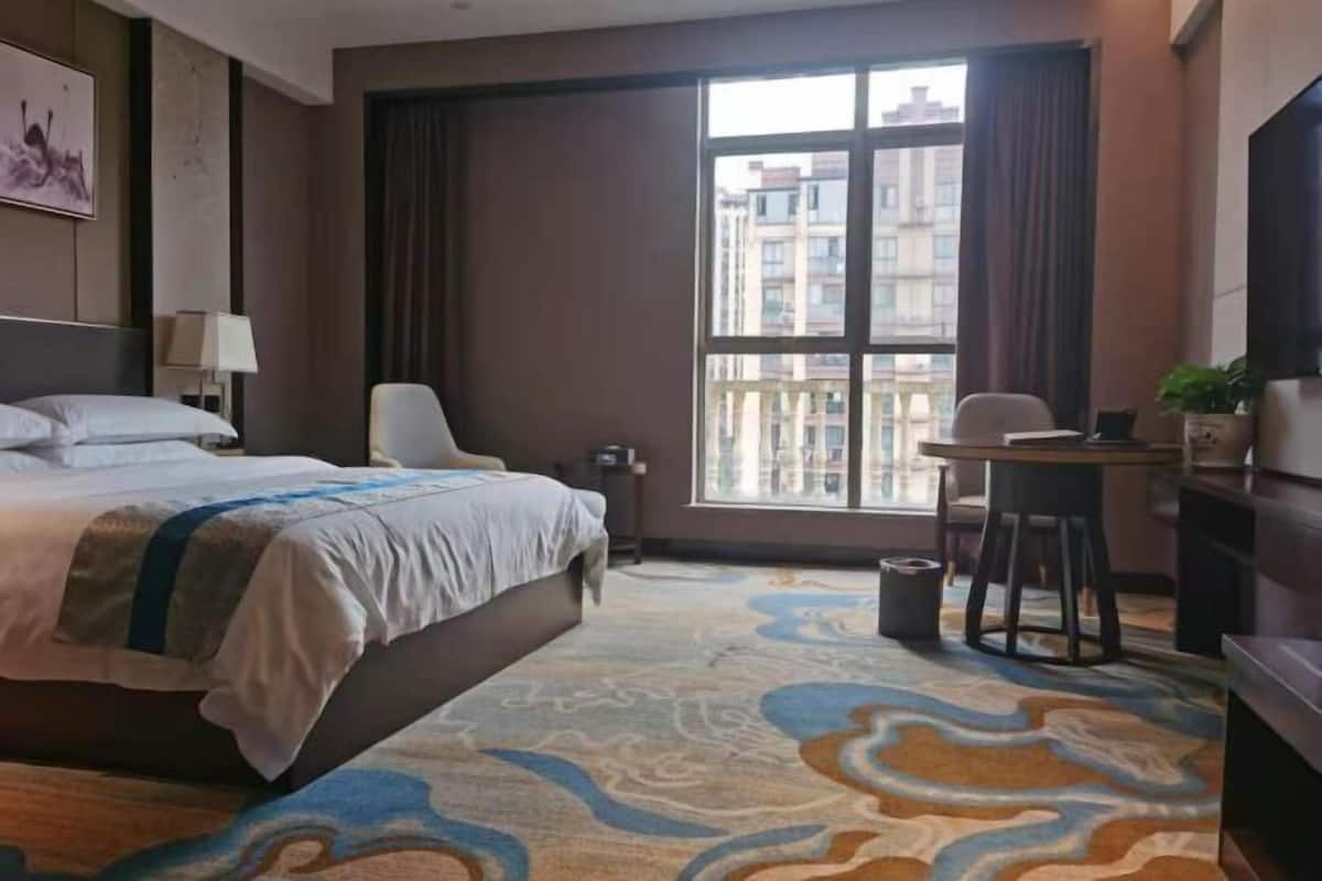 Une chambre d’hôtel équipée d’un lit, d’un bureau, d’une chaise, d’une télévision et d’une grande fenêtre donnant sur des immeubles.