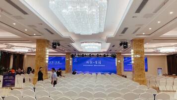 A conference room with rows of white chairs, a stage with a blue backdrop, and a large chandelier.