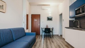 A compact living space with a sofa, a dining table with chairs, a kitchen area with cabinets and a microwave, and a door leading to another room. - Al 13 Blu 2 - Lakeside L&b (Stresa)