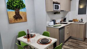 Una cocina compacta con microondas, lavaplatos y armarios de madera. También se ve una mesa de comedor con sillas verdes y un cuadro de un árbol.