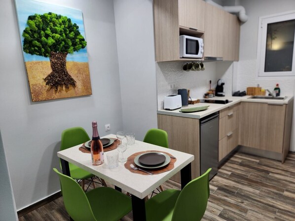 A compact kitchen with a microwave, sink, and wooden cabinets. A dining table with green chairs and a painting of a tree are also visible. - Central area of Athens, two bedroom Cozy Apartment with garden and terrace (Athina)